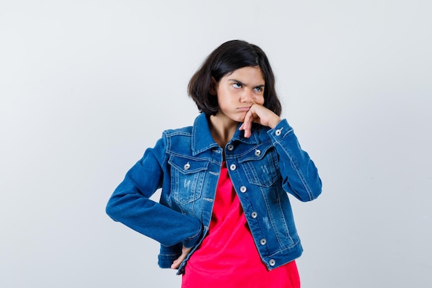 Giovane ragazza in piedi in posa di pensiero in maglietta rossa e giacca di jeans e guardando pensieroso, vista frontale.