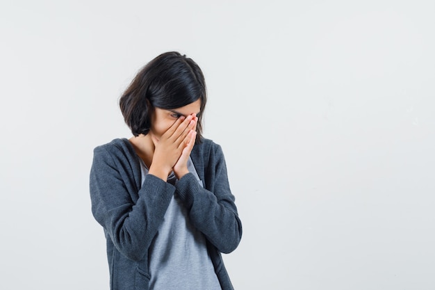 Giovane ragazza in piedi in posa di preghiera in maglietta grigio chiaro e felpa con cappuccio con zip grigio scuro e sguardo concentrato.