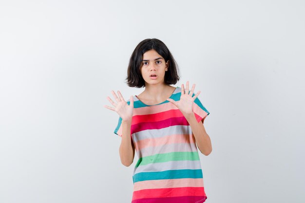 Giovane ragazza in t-shirt a righe colorate alzando i palmi in gesto di resa e guardando scioccata, vista frontale.