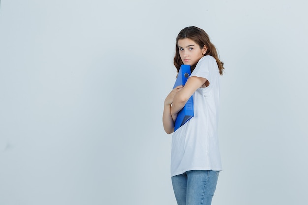 Giovane ragazza in t-shirt bianca che abbraccia cartella e guardando imbarazzato, vista frontale.
