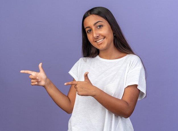 Giovane ragazza in t-shirt bianca che guarda l'obbiettivo con un sorriso sulla faccia felice che punta con il dito indice al lato in piedi su sfondo blu