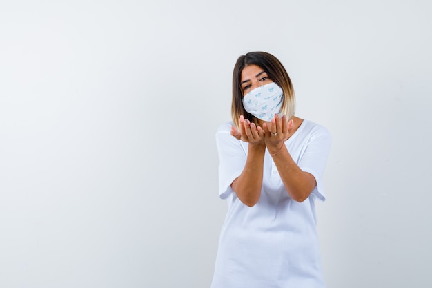 Giovane ragazza in t-shirt bianca e maschera che allunga le mani a coppa e sembra allegro, vista frontale.