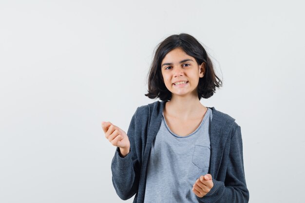 Giovane ragazza in t-shirt grigio chiaro e felpa con cappuccio con zip grigio scuro che stringe i pugni e sembra carina