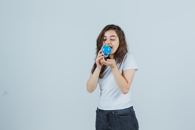 Giovane ragazza in t-shirt, jeans cercando di mordere il globo e guardando pazzo, vista frontale.