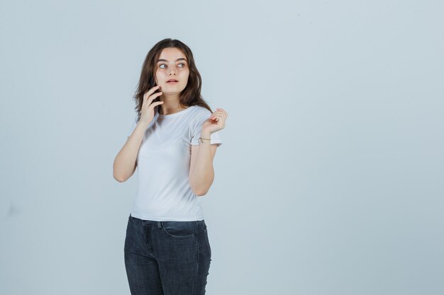 Giovane ragazza in t-shirt, jeans parlando al cellulare mentre guarda lontano e guardando serio, vista frontale.