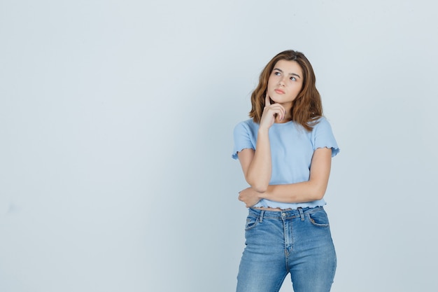 Giovane ragazza in t-shirt, jeans tenendo il dito sul mento e guardando pensieroso, vista frontale.