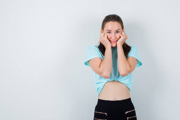 Giovane ragazza in t-shirt turchese, pantaloni appoggiati sulle guance sulle mani e dall'aspetto carino, vista frontale.