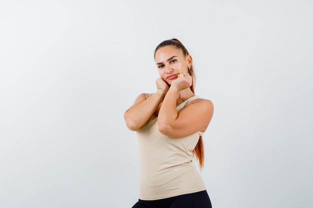 Giovane ragazza in top beige, pantaloni neri che coprono la faccia sulle mani e che sembra carina, vista frontale