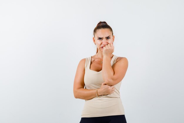 Giovane ragazza in top beige, pantaloni neri che morde i pugni e sembra eccitata, vista frontale.