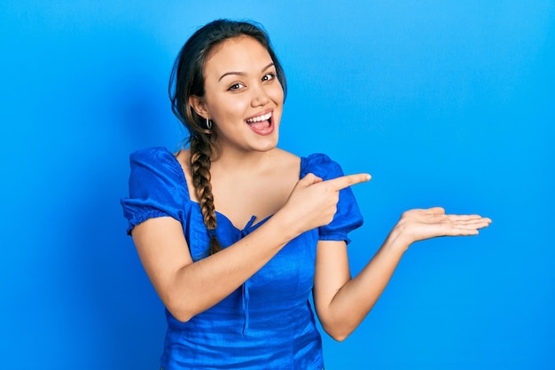 Giovane ragazza ispanica che indossa abiti casual stupita e sorridente alla telecamera mentre si presenta con la mano e punta con il dito