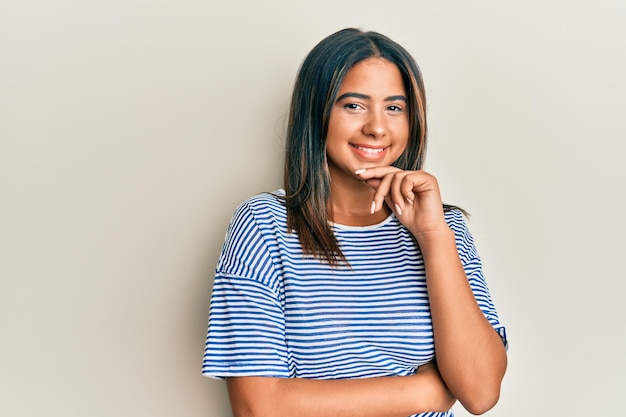 Giovane ragazza latina che indossa abiti casual sorridente guardando fiducioso la fotocamera con le braccia incrociate e la mano sul mento. pensare positivo.