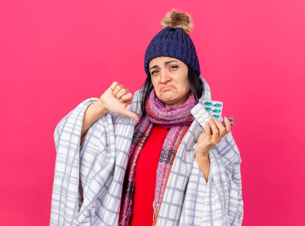 Giovane ragazza malata caucasica dispiaciuta che indossa cappello invernale e sciarpa avvolti in confezioni di plaid che tengono le pillole mediche che mostrano il pollice verso il basso isolato sul muro cremisi