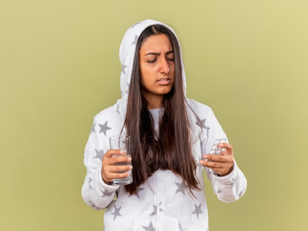 Giovane ragazza malata di pensiero che mette sul cappuccio che tiene il bicchiere d'acqua e guardando le pillole in mano isolato su sfondo verde oliva