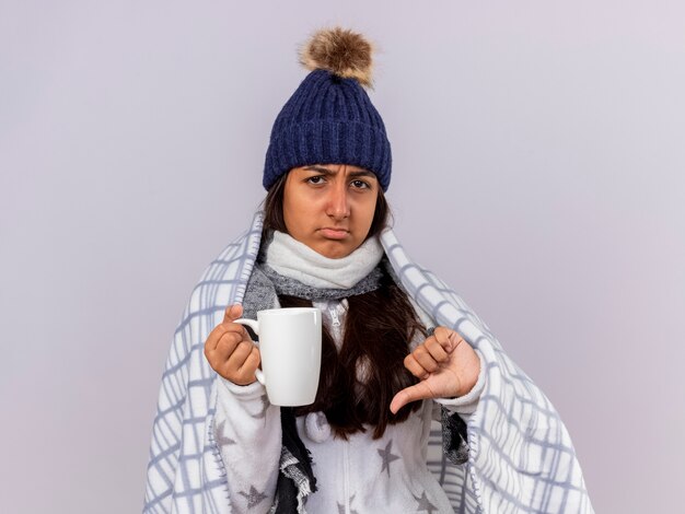 Giovane ragazza malata dispiaciuta che indossa il cappello invernale con sciarpa avvolta in una tazza di tè che tiene plaid che mostra il pollice verso il basso