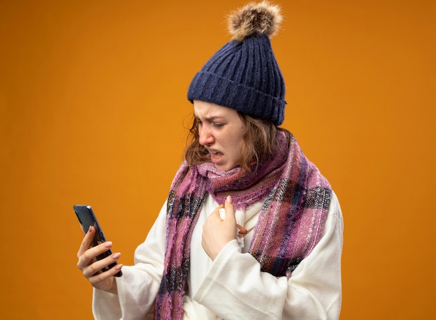 Giovane ragazza malata dispiaciuta che indossa una veste bianca e cappello invernale con la sciarpa che tiene e guardando i punti del telefono a se stessa isolata sulla parete arancione