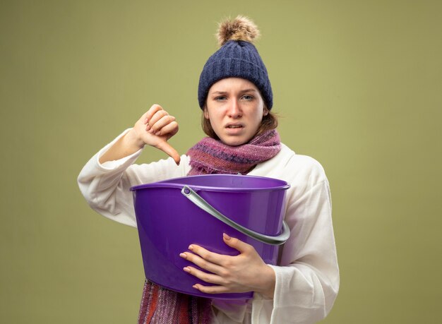 Giovane ragazza malata dispiaciuta che indossa veste bianca e cappello invernale con sciarpa che tiene secchio di plastica che mostra il pollice verso il basso isolato su verde oliva