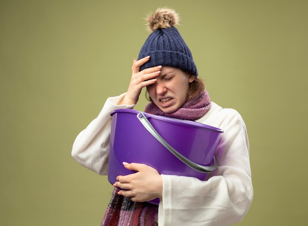 Giovane ragazza malata dolorante che indossa una veste bianca e cappello invernale con la sciarpa che tiene il secchio di plastica che mette la mano sulla fronte isolata su verde oliva