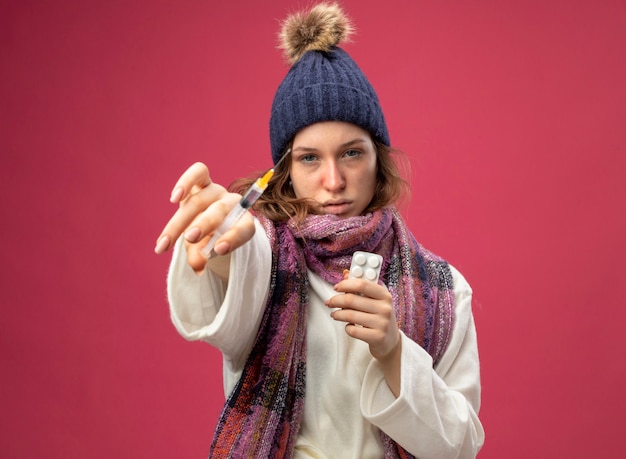 Giovane ragazza malata guardando dritto davanti a sé indossando una veste bianca e cappello invernale con sciarpa tenendo le pillole e tenendo la siringa isolata sul rosa