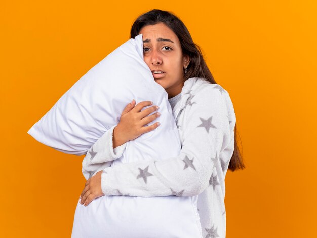 Giovane ragazza malata interessata che guarda l'obbiettivo che abbraccia il cuscino isolato su priorità bassa gialla