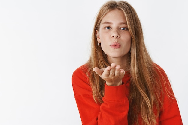 giovane ragazza mantica con capelli biondi, lentiggini e occhi azzurri che piegano le labbra tenendo i palmi vicino alla bocca, inviando un bacio d'aria con caldi sentimenti d'amore su grigio.