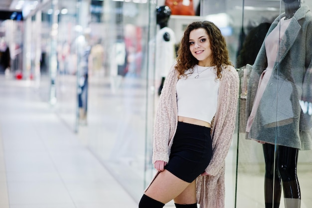 Giovane ragazza modella riccia posata su minigonna al grande centro commerciale vicino alla vetrina con manichino