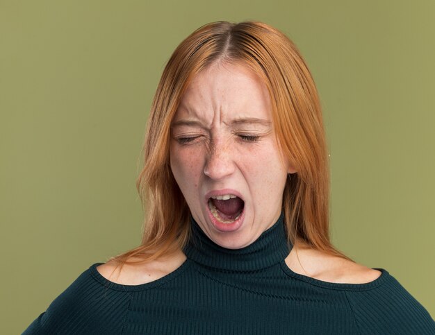 Giovane ragazza rossa infastidita dello zenzero con le lentiggini che stanno con gli occhi chiusi isolati sulla parete verde oliva con lo spazio della copia