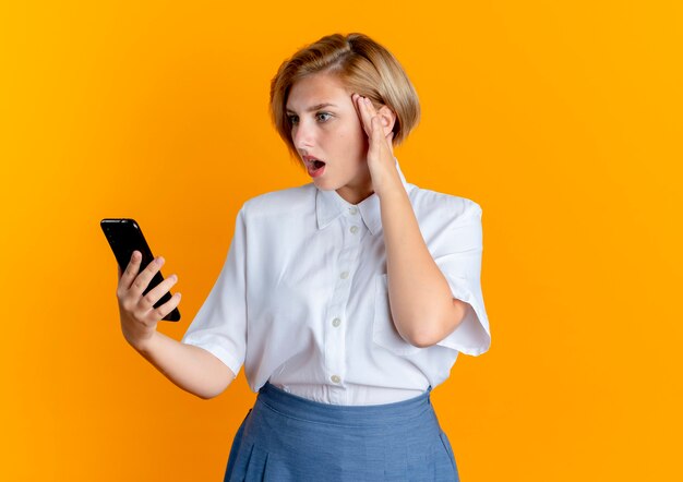 Giovane ragazza russa bionda sorpresa mette la mano sulla testa tenendo e guardando il telefono isolato su sfondo arancione con spazio di copia