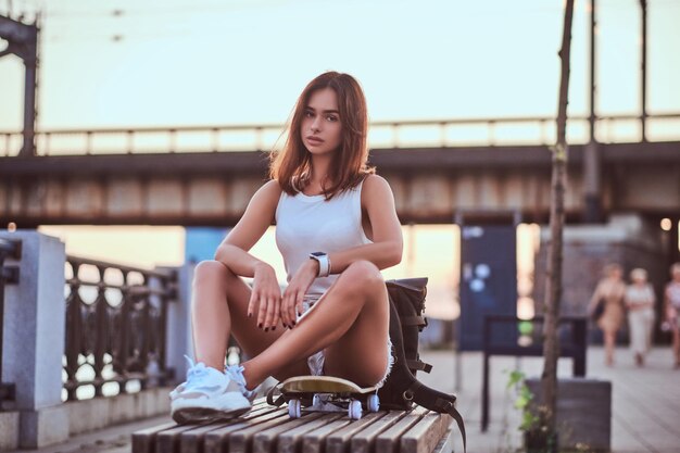 Giovane ragazza sensuale skater vestita in pantaloncini corti e t-shirt seduta sull'argine durante un bel tramonto.