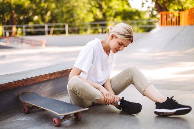 Giovane ragazza skater che tiene la sua gamba dolorosa con lo skateboard vicino al moderno skatepark