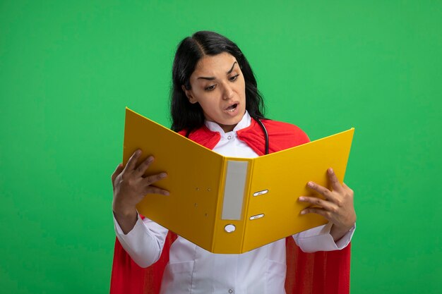 Giovane ragazza sorpresa del supereroe che indossa la veste medica con lo stetoscopio che tiene e che esamina la cartella isolata sul verde