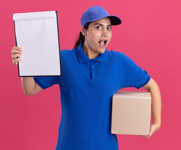 Giovane ragazza sorpresa di consegna che indossa l'uniforme con la scatola della tenuta del cappuccio con la lavagna per appunti isolata sulla parete rosa