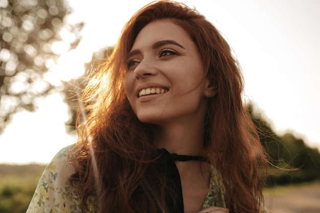 Giovane ragazza sorridente con acconciatura rossa e benda nera sul collo in abito estivo moderno sorridente e distogliendo lo sguardo all'aperto