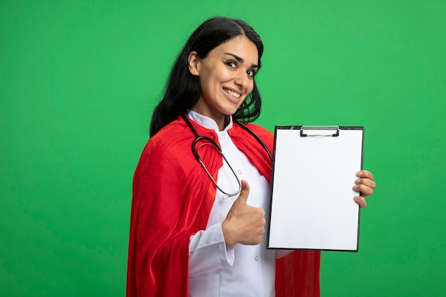 Giovane ragazza sorridente del supereroe che indossa la veste medica con lo stetoscopio che tiene appunti che mostra il pollice in su isolato sul verde