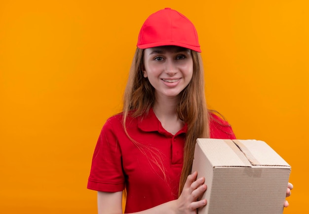 Giovane ragazza sorridente di consegna in scatola di tenuta uniforme rossa sullo spazio arancione isolato con lo spazio della copia