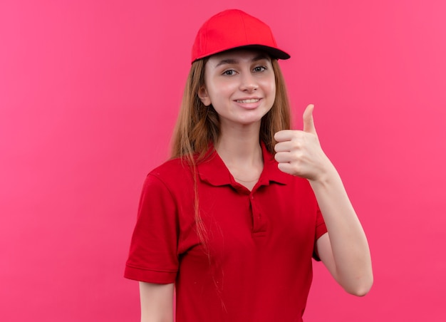 Giovane ragazza sorridente di consegna in uniforme rossa che mostra pollice in su sulla parete rosa isolata con lo spazio della copia