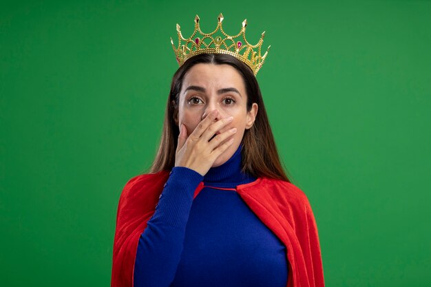 Giovane ragazza spaventata del supereroe che indossa la bocca coperta della corona con la mano isolata sul verde