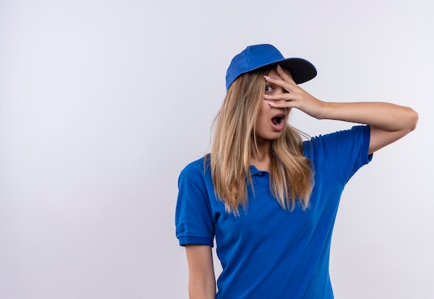 Giovane ragazza spaventata delle consegne che indossa l'uniforme blu e il cappuccio coperto gli occhi con la mano isolata sulla parete bianca con lo spazio della copia