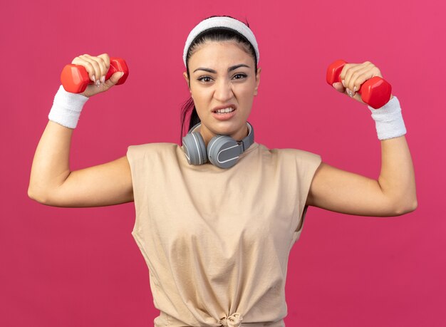 Giovane ragazza sportiva caucasica tesa che indossa fascia e braccialetti con le cuffie intorno al collo guardando i manubri di sollevamento anteriori isolati sulla parete rosa