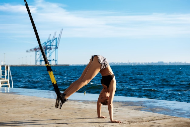 Giovane ragazza sportiva che si allena con trx vicino al mare al mattino.