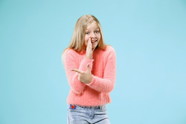 Giovane ragazza teenager che bisbiglia un segreto dietro la sua mano isolata sulla parete blu alla moda dello studio