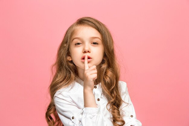 Giovane ragazza teenager che bisbiglia un segreto dietro la sua mano isolata sulla parete rosa alla moda dello studio.