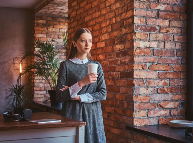 Giovane ragazza vestita con un elegante abito grigio tiene una tazza di caffè da asporto distogliendo lo sguardo mentre si trova in una stanza con interni soppalcati.