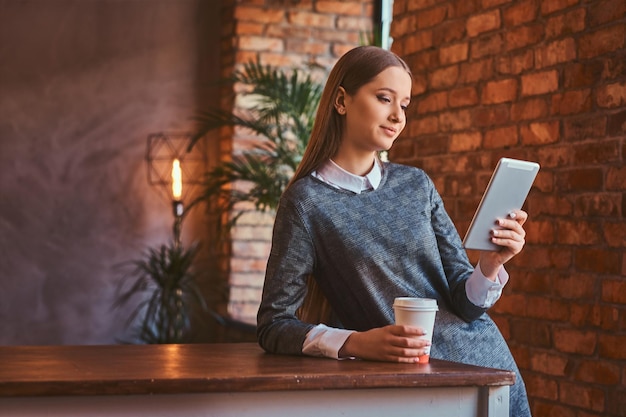 Giovane ragazza vestita con un elegante abito grigio tiene una tazza di caffè da asporto e utilizza un tablet mentre si appoggia sul tavolo in una stanza con interni soppalcati.