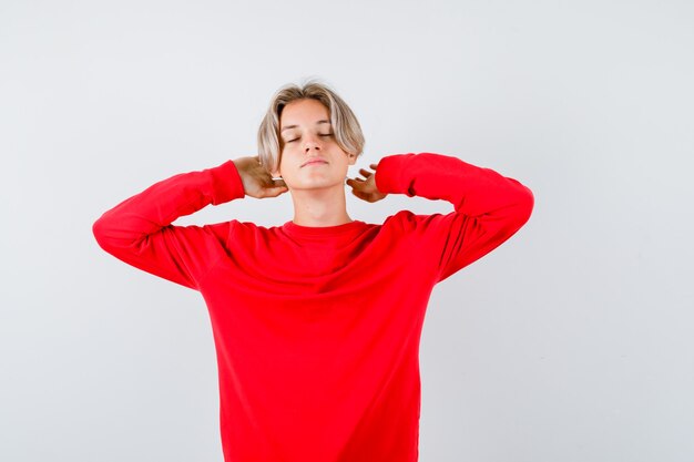 Giovane ragazzo adolescente che allunga le braccia dietro la testa, chiude gli occhi con un maglione rosso e sembra rilassato. vista frontale.