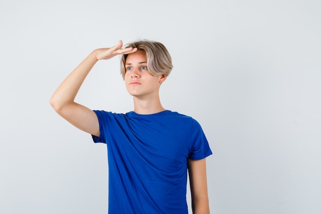 Giovane ragazzo adolescente che guarda lontano con la mano sopra la testa in maglietta blu e sembra concentrato, vista frontale.