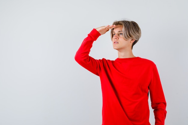 Giovane ragazzo adolescente che guarda lontano con la mano sopra la testa in un maglione rosso e sembra concentrato, vista frontale.