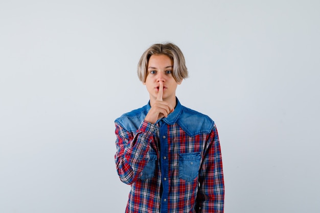 Giovane ragazzo adolescente che mostra gesto di silenzio in camicia a quadri e guarda attento. vista frontale.