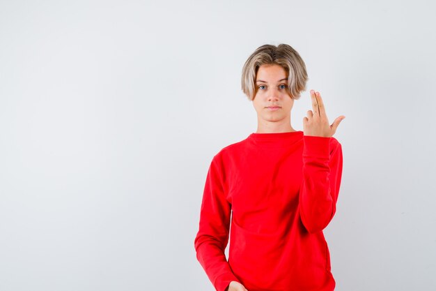 Giovane ragazzo adolescente che mostra il gesto della pistola in maglione rosso e sembra serio, vista frontale.