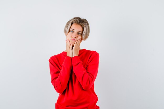 Giovane ragazzo adolescente con le mani sulle guance in maglione rosso e dall'aspetto triste. vista frontale.