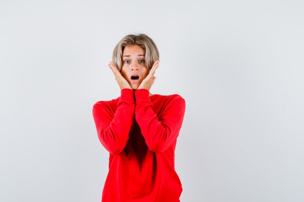 Giovane ragazzo adolescente con le mani sulle guance in maglione rosso e sembra agitato. vista frontale.